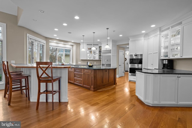 kitchen with pendant lighting, a kitchen island with sink, white cabinetry, stainless steel double oven, and light wood-type flooring