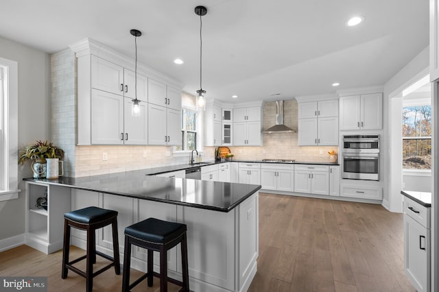 kitchen with a peninsula, white cabinets, wall chimney exhaust hood, dark countertops, and decorative light fixtures