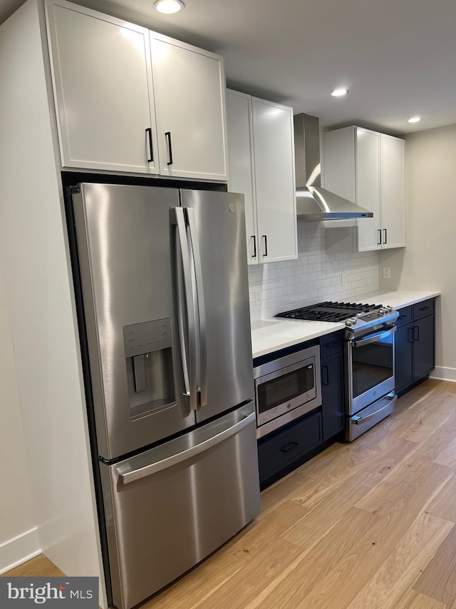 kitchen with backsplash, wall chimney exhaust hood, light wood-type flooring, appliances with stainless steel finishes, and white cabinetry