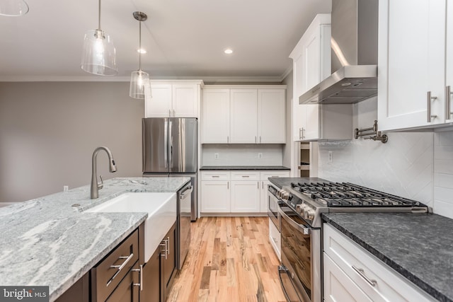 kitchen featuring light wood-type flooring, wall chimney exhaust hood, stainless steel appliances, dark stone countertops, and white cabinets