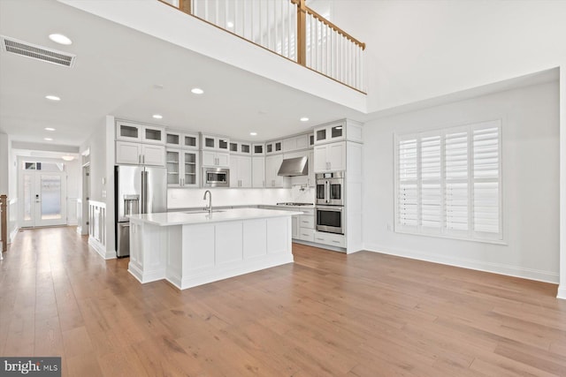 kitchen with wall chimney exhaust hood, stainless steel appliances, light hardwood / wood-style floors, a center island with sink, and white cabinets