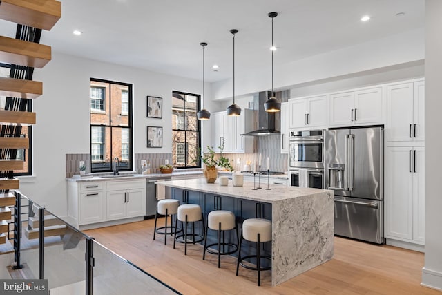 kitchen featuring a center island, wall chimney range hood, hanging light fixtures, white cabinetry, and stainless steel appliances