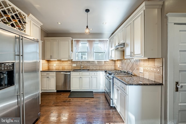 kitchen featuring sink, dark wood-type flooring, dark stone countertops, decorative light fixtures, and appliances with stainless steel finishes