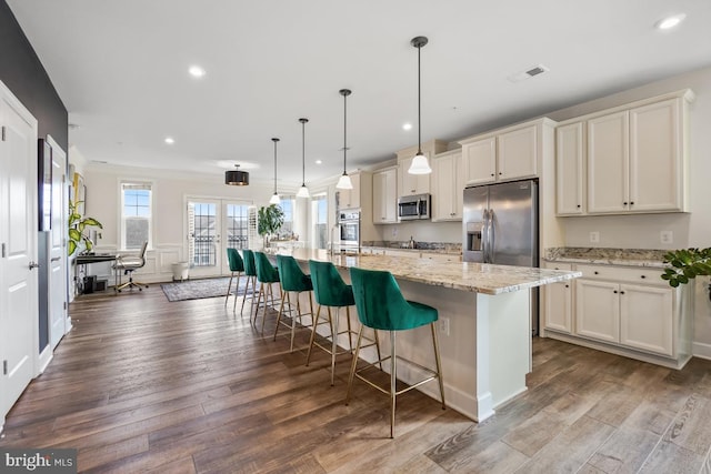 kitchen with light stone countertops, appliances with stainless steel finishes, pendant lighting, hardwood / wood-style flooring, and an island with sink