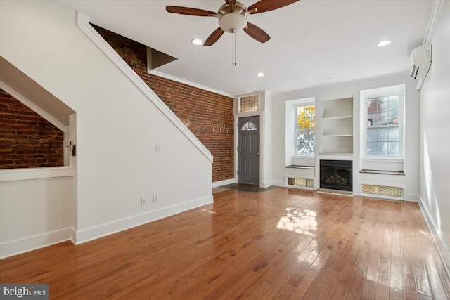 unfurnished living room featuring a wall mounted air conditioner, brick wall, built in shelves, ceiling fan, and wood-type flooring