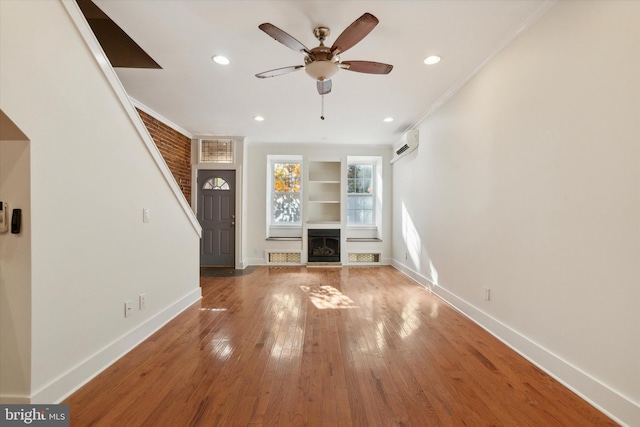 unfurnished living room featuring ceiling fan, built in features, an AC wall unit, crown molding, and wood-type flooring