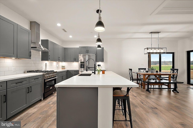 kitchen with gray cabinetry, stainless steel appliances, a kitchen island with sink, wall chimney range hood, and light hardwood / wood-style flooring