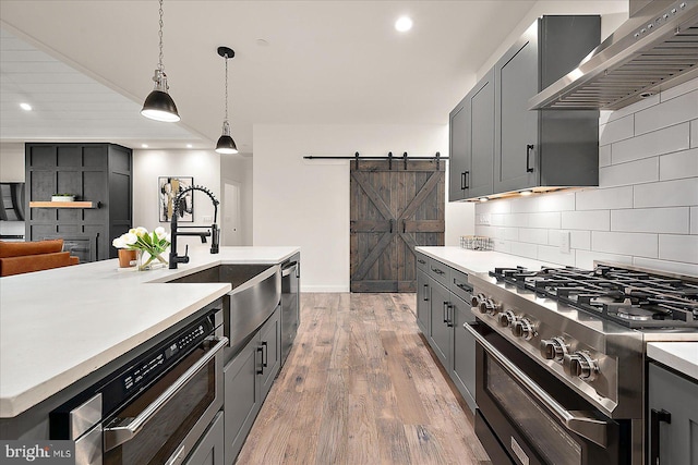 kitchen featuring wall chimney range hood, hanging light fixtures, light hardwood / wood-style flooring, a barn door, and appliances with stainless steel finishes