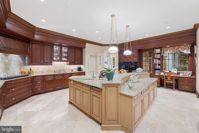 kitchen with backsplash, crown molding, hanging light fixtures, light stone countertops, and an island with sink