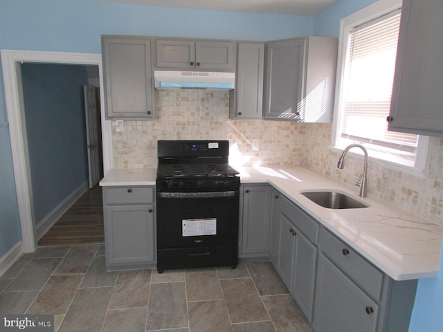 kitchen with decorative backsplash, gray cabinetry, black gas stove, a sink, and under cabinet range hood