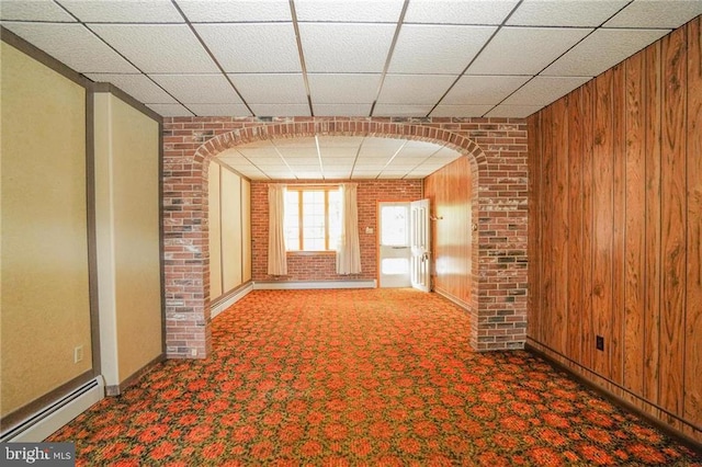 carpeted spare room featuring a paneled ceiling, baseboard heating, wooden walls, and brick wall