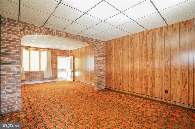 unfurnished living room with a paneled ceiling, wooden walls, and brick wall