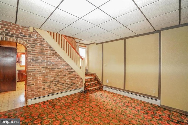 basement featuring a drop ceiling, a baseboard radiator, and brick wall