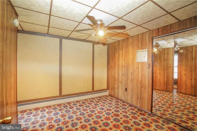 basement with baseboard heating, ceiling fan, wooden walls, and a drop ceiling