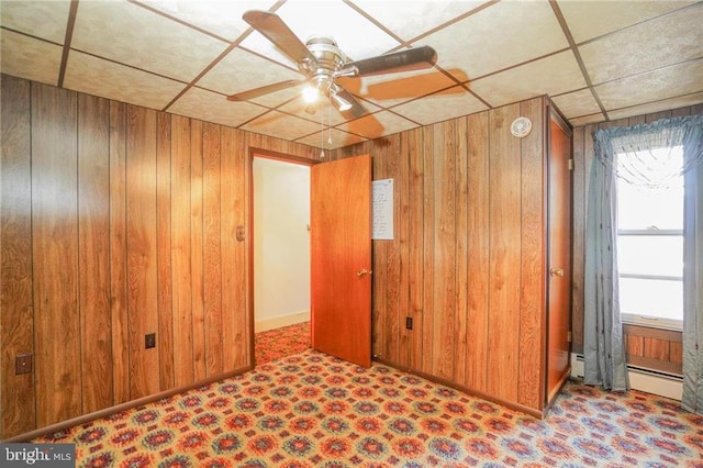 empty room featuring a paneled ceiling, ceiling fan, wooden walls, and a baseboard radiator