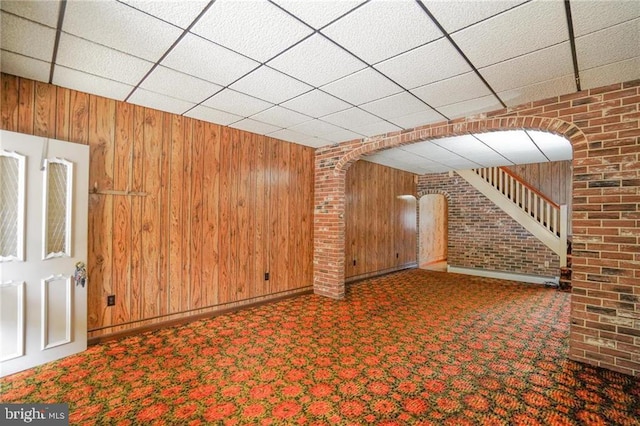 basement featuring wooden walls, a drop ceiling, carpet floors, and brick wall