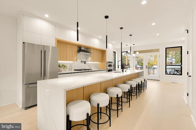 kitchen featuring a large island with sink, wall chimney range hood, hanging light fixtures, white cabinetry, and stainless steel appliances