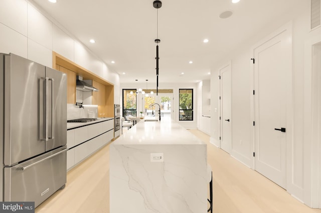 kitchen with appliances with stainless steel finishes, a kitchen island with sink, wall chimney range hood, pendant lighting, and white cabinets