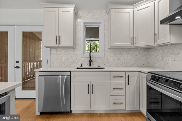 kitchen with appliances with stainless steel finishes, light wood-type flooring, wall chimney range hood, sink, and white cabinetry