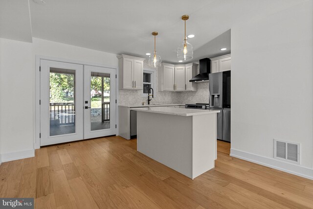 kitchen with wall chimney range hood, appliances with stainless steel finishes, decorative light fixtures, a kitchen island, and white cabinetry