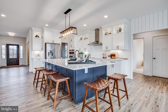 kitchen with hanging light fixtures, wall chimney exhaust hood, an island with sink, white cabinetry, and stainless steel appliances