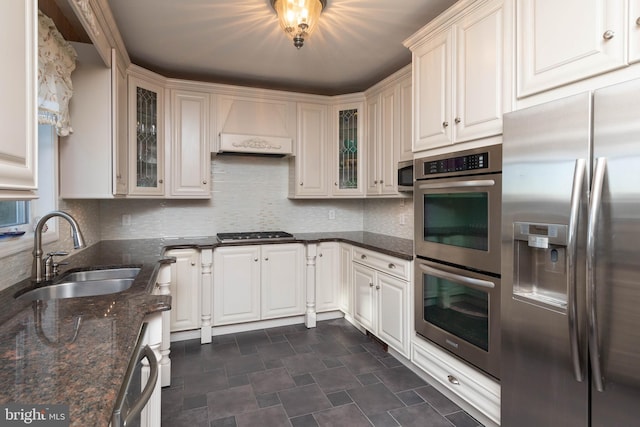 kitchen with decorative backsplash, custom exhaust hood, stainless steel appliances, sink, and dark stone countertops