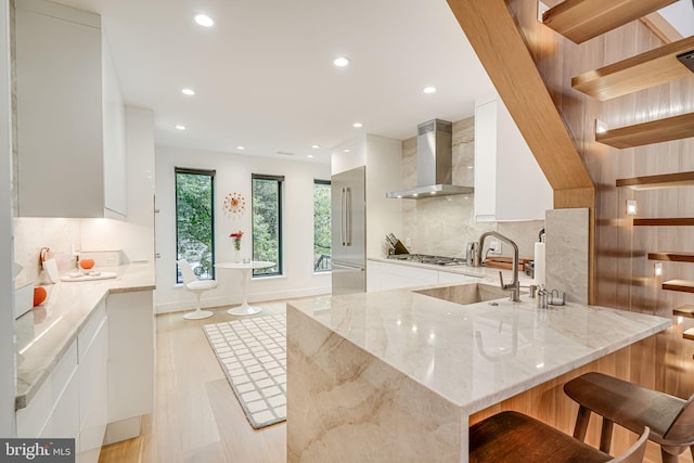 kitchen with backsplash, a kitchen bar, light stone countertops, white cabinets, and wall chimney exhaust hood