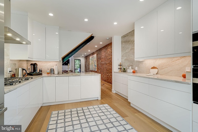 kitchen with brick wall, white cabinets, wall chimney range hood, stainless steel gas cooktop, and light wood-type flooring