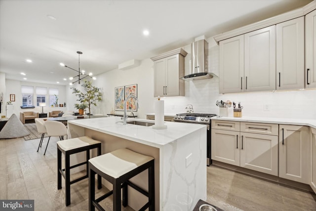 kitchen with wall chimney range hood, hanging light fixtures, gas range, an island with sink, and light hardwood / wood-style floors