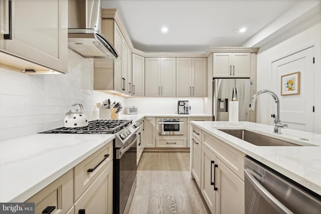 kitchen with sink, wall chimney exhaust hood, light stone countertops, light hardwood / wood-style floors, and stainless steel appliances