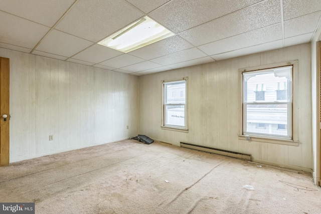 carpeted empty room with a drop ceiling, wood walls, and a baseboard heating unit