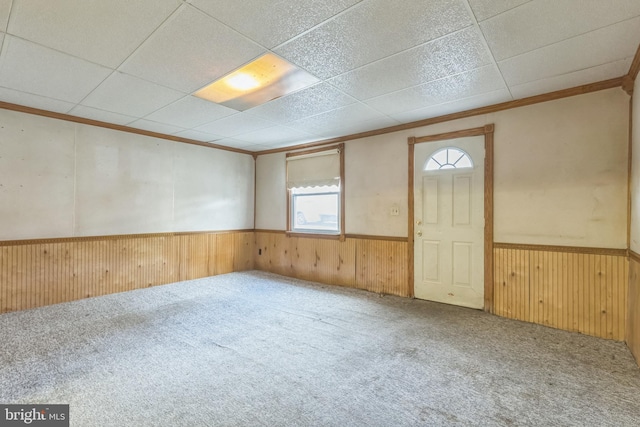 carpeted empty room with a drop ceiling, wood walls, and crown molding