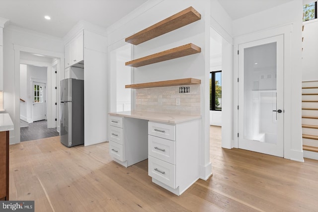 kitchen with stainless steel fridge, backsplash, built in desk, white cabinets, and light hardwood / wood-style floors