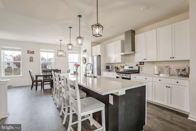 kitchen featuring white cabinetry, wall chimney exhaust hood, hanging light fixtures, a kitchen island with sink, and appliances with stainless steel finishes