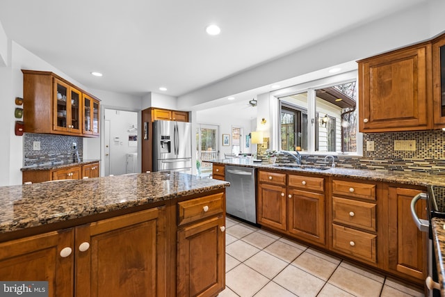 kitchen featuring brown cabinetry, glass insert cabinets, dark stone countertops, stainless steel appliances, and a sink