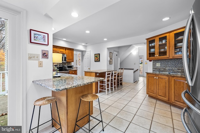 kitchen featuring light tile patterned flooring, appliances with stainless steel finishes, a center island, brown cabinetry, and a kitchen bar