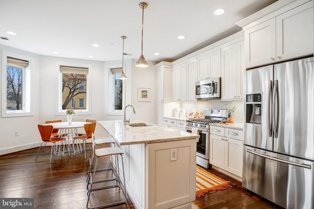 kitchen featuring an island with sink, light stone counters, stainless steel appliances, pendant lighting, and a sink