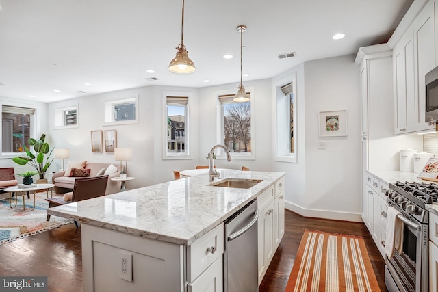 kitchen with a kitchen island with sink, a sink, appliances with stainless steel finishes, light stone countertops, and decorative light fixtures