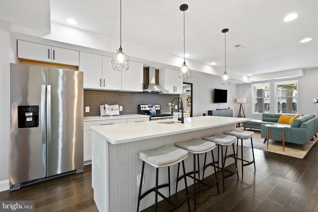 kitchen featuring white cabinets, wall chimney range hood, hanging light fixtures, an island with sink, and appliances with stainless steel finishes