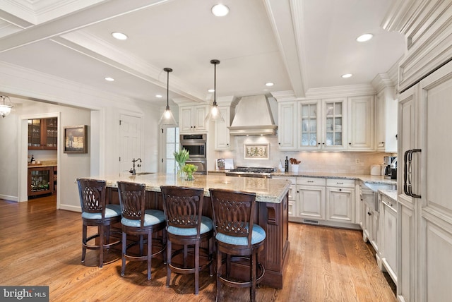 kitchen featuring a kitchen island with sink, wood finished floors, appliances with stainless steel finishes, custom exhaust hood, and beamed ceiling