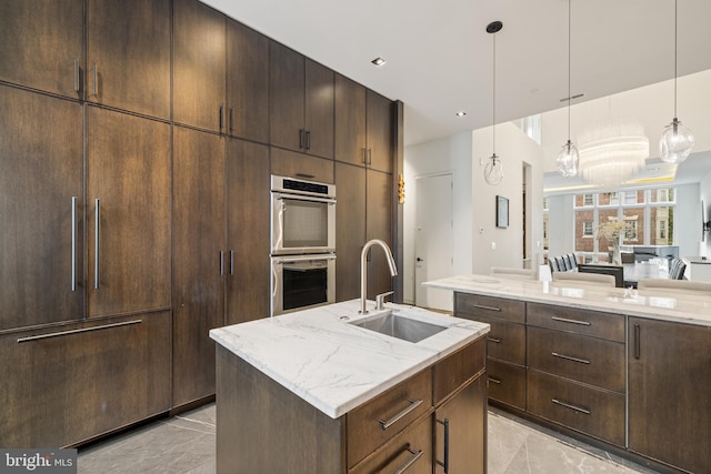 kitchen featuring a kitchen island with sink, hanging light fixtures, sink, double oven, and light stone counters