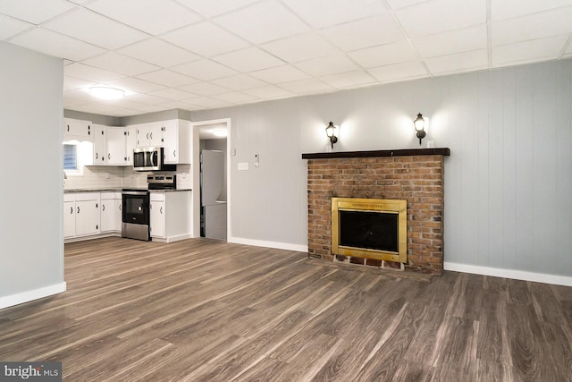 unfurnished living room featuring a paneled ceiling, wood walls, dark wood-type flooring, and a brick fireplace