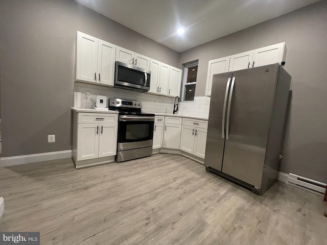 kitchen with backsplash, white cabinets, baseboard heating, appliances with stainless steel finishes, and light hardwood / wood-style floors