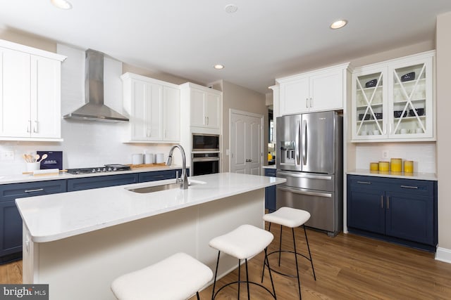 kitchen with appliances with stainless steel finishes, a center island with sink, blue cabinets, and wall chimney exhaust hood