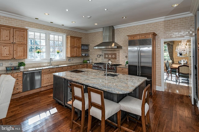 kitchen featuring light stone counters, stainless steel appliances, dark wood-type flooring, wall chimney range hood, and a center island with sink