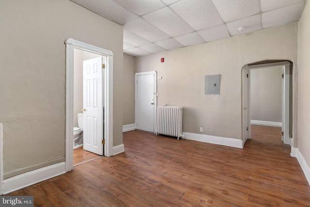 unfurnished bedroom featuring a drop ceiling, electric panel, hardwood / wood-style flooring, connected bathroom, and radiator heating unit