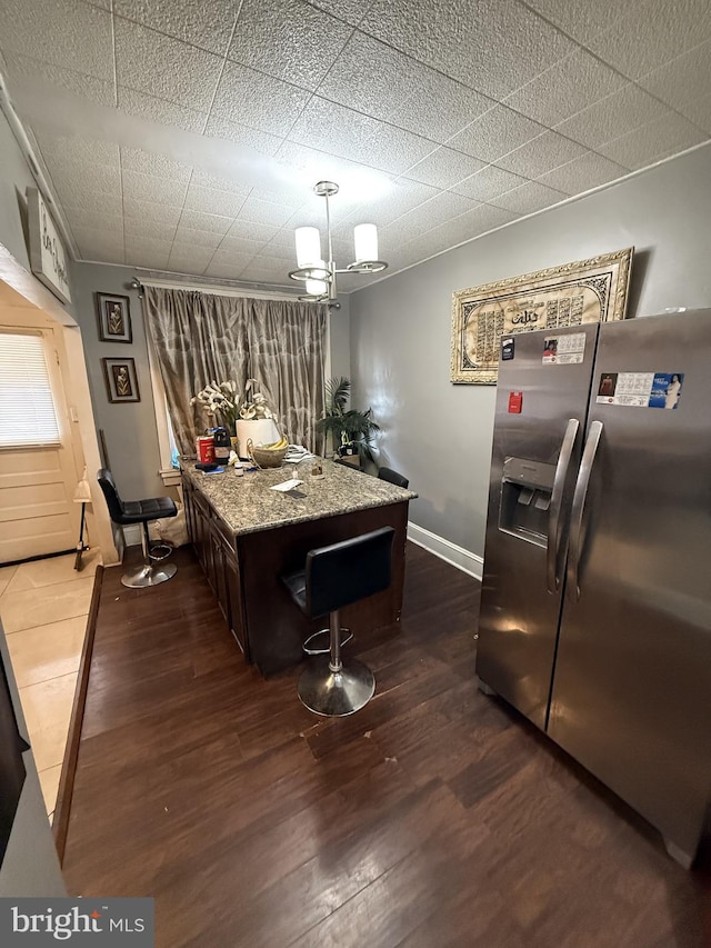 kitchen featuring light stone countertops, a kitchen bar, dark brown cabinetry, dark wood-type flooring, and stainless steel fridge with ice dispenser