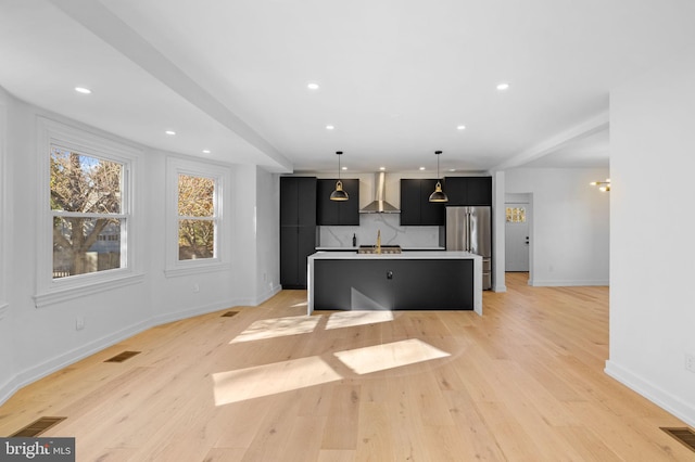 kitchen with wall chimney range hood, light hardwood / wood-style flooring, stainless steel refrigerator, hanging light fixtures, and an island with sink