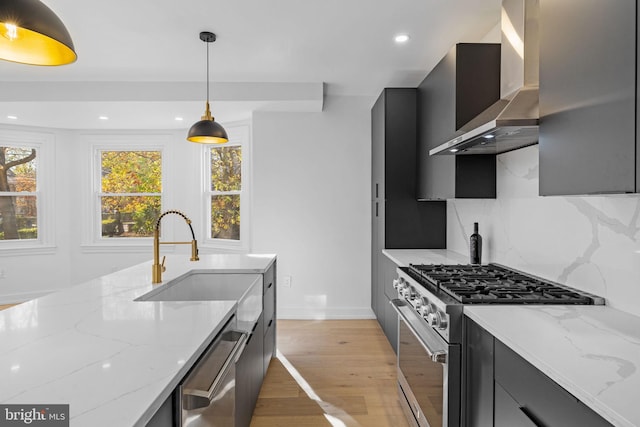 kitchen with light stone counters, stainless steel appliances, wall chimney range hood, pendant lighting, and light hardwood / wood-style flooring