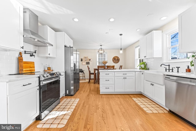 kitchen with wall chimney exhaust hood, stainless steel appliances, decorative light fixtures, light hardwood / wood-style flooring, and white cabinets